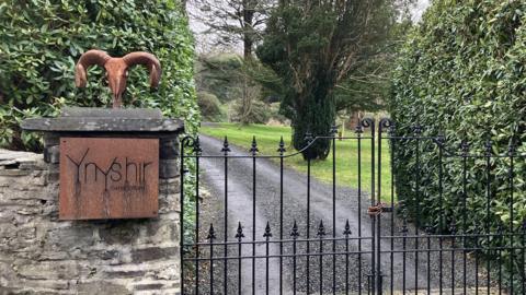 The gated entrance to Ynshir restaurant. A black cast iron gate leads up a drive. On the wall is a bronze coloured sign with the restaurant's name on it. 