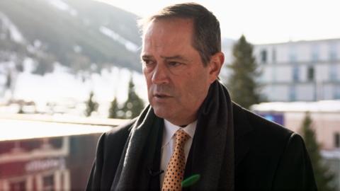 Cisco boss Chuck Robbins wearing a dark suit and scarf over a yellow patterned tie. He stands with snowy mountains in the background.