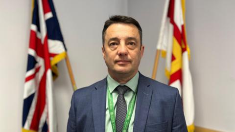 A man in a suit standing in front of a union flag and a Guernsey flag.