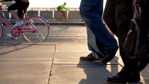 Pedestrians walk together along the side of a road, with their feet and legs visible. Two cyclists can be seen riding bikes in the background.