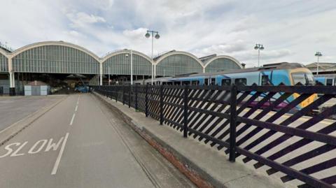 A blue train with a yellow cab outside Paragon Station, Hull. The station features five large, partly-glazed arches, which support the roof. To the left is a roadway, which is separated from the platform by a wooden, slatted fence.