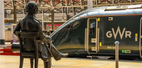 A green train pulled into the station with the white letters GWR on it. There is a metallic statue of a man sitting down facing the train on the platform with his legs crossed.