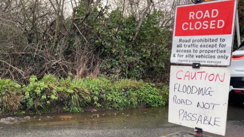 A flooded road in Guernsey with a hand written sign reading Caution flooding road not passable. A formal sign from Traffic and Highways reads Road Closed - Road prohibited to all traffic except for access to properties and for site vehicles only.