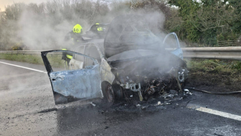 The picture shows a badly burned car on the side of a road. The front of the vehicle is almost completely destroyed, with the engine area exposed and charred. Thick smoke is rising from the wreckage, suggesting the fire has only recently been put out. The car doors are open, and the paintwork is scorched and peeling. Two firefighters wearing high-visibility helmets and protective gear are standing behind the car, partially obscured by the smoke.