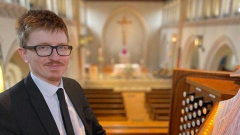 A man with ginger hair and black glasses smiles at the camera. He is wearing a black suit and tie with a white shirt. The top of an organ is seen in the picture and the background of the image, which has been blurred to focus on the man, is that of a large white church with brown palls.