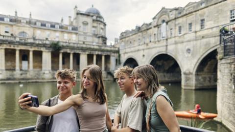 A group of young tourists taking a selfie in front of Bath's waterways and tunnels on an overcast day.