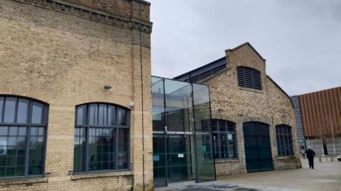 Sand Martin House in Peterborough. It is a listed former Victorian brick railway building containing two locomotive sheds transformed into offices. In between the buildings is is a modern black framed glass entrance and atrium, linking them. On the right a man in black can be seen walking away from the building. 