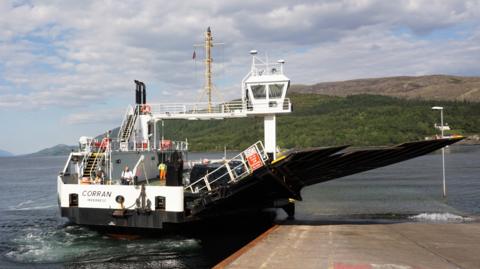The Corran Ferry's main vessel, MV Corran, is next to a slipway. The ferry's propellers churn Loch Linnhe's waters. The opposite shore, with tree-covered hills, is in the background.