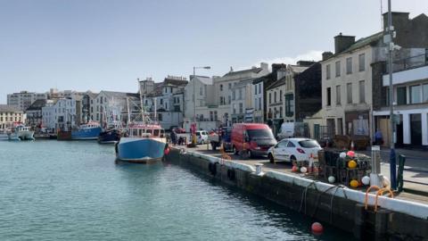 A row of boats line a working harbour in Ramsey, where cars are parked along the road near to the boats on the left. To the right is a row of different shaped and heights of terrace houses, shops, and public houses. The tide is fairly high on a sunny day.