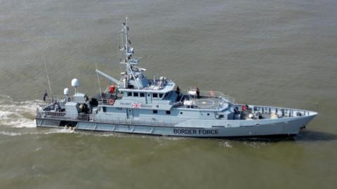 Large grey boat in the sea with Border Force painted on the side. It also has a Union Jack on the side next to its name Valiant. There is equipment on board and a helipad.
