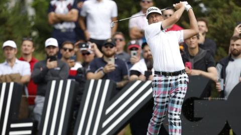 Ian Poulter in the middle of a follow-through after hitting a shot during a professional tournament. In the background are large black signs featuring bold white striped graphics.