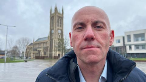 A man with a shaven head stands in a town square in front of a Victorian church, built in stone and brown brick, with a tall tower. He is wearing a dark blue coat and light blue shirt.