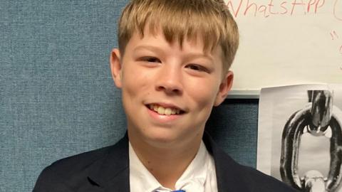 Theo smiles at the camera, he is wearing a school uniform which consists of a black blazer, white shirt and bright blue tie.