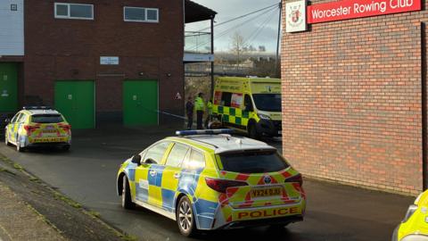 Two police cars and an ambulance are parked outside a red brick building with a sign that reads: "Worcester Rowing Club." 
A couple emergency workers in high viz clothing are seen standing by the ambulance.