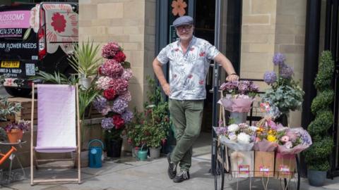Image shows a man wearing green trousers and a floral shirt, leaning on a trolly of a display of flowers. To the left of the picture is a separate display of purple and pink flowers and a purple deck chair.