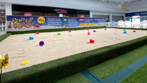 A large section in a shopping centre which is filled with sand. On the sand are multicoloured buckets and spades. Around the perimeter is fake grass. Behind the giant sandpit are shops.