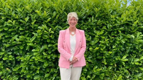 Woman wearing a pink jacket, white trousers and top with a green bush in the background