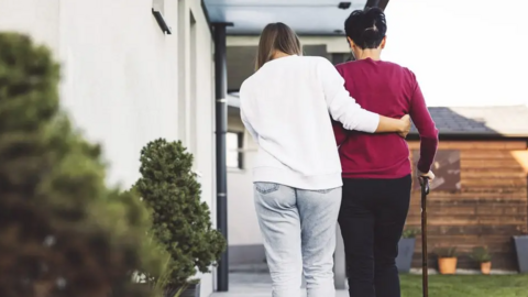 A woman assists another, leaning on a walking stick, as they walk together past a house