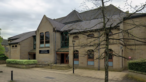 A stone building with arched windows and a central entrance marked “Harrogate Justice Centre”, with steps, railings, trees and greenery in front under an overcast sky.