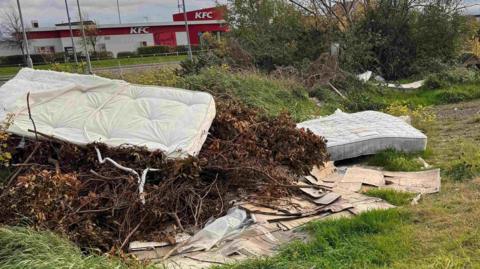 Multiple mattresses are piled on to sticks, plastic bags and wet cardboard in a field. A KFC building can be seen in the distance.