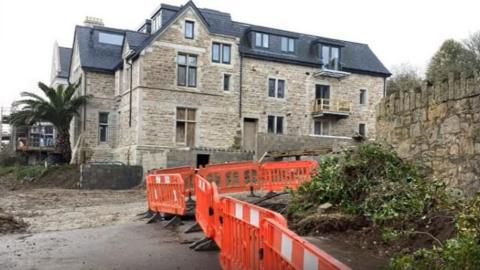 A large four-storey building has safety barriers to one side. There is a palm tree in front of the property.