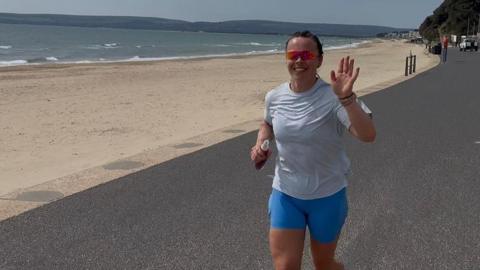 Tegan Pryce is waving to the camera as she runs along a road which runs parallel to an empty beach on a sunny day. She is wearing pale blue running shorts, a white T-shirt and a pair of sunglasses.