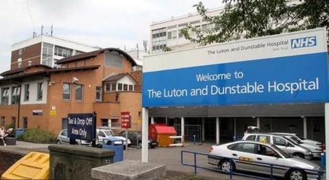 The front of the  hospital with a large blue/white sign saying "Welcome to the Luton and Dunstable Hospital". Taxis are parked behind it and there are large hospital buildings in the background. 