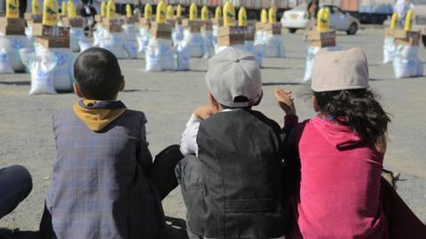 Three children sitting, with their backs to the camera, facing bags of aid
