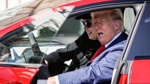 Donald Trump and Elon Musk sit inside a red Tesla; Musk smiles and looks ahead while Trump speaks