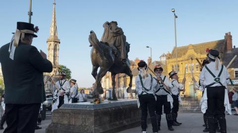 Morris dancers dancing around the Fine Lady Statue. One of them is taking pictures. It is a sunny day.