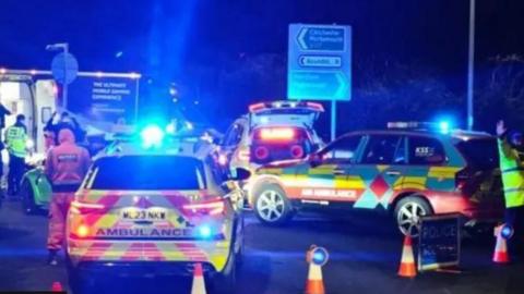 A night-time shot showing ambulance vehicles and emergency services personnel in high vis on a Worthing road coned off with a sign in the background showing Chichester and Arundel