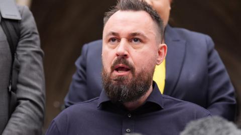 Martin Hibbert, dressed in a blue shirt, speaking in front of microphones following a High Court appearance
