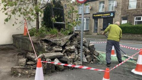 A workman clears up at the crash scene where a smashed wall is sealed off with red and white tape and orange and white traffic cones.