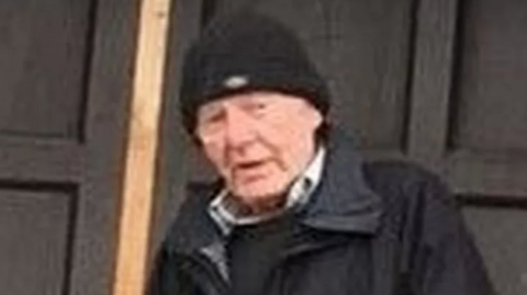 An image of Alan Lloyd. It is a blurred image, possibly from a distance and zoomed in. He is a white man, elderly, wearing a black beanie hat, black rain coat, and black jumper with a white or light-coloured shirt collar showing. He has a neutral expression.