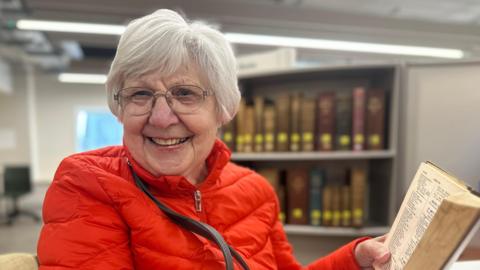 Valerie Henry is sitting on a chair on the left. She has short grey hair and is wearing a red coat, glasses, and is holding a book on the right. Behind her is a small bookcase with old books.