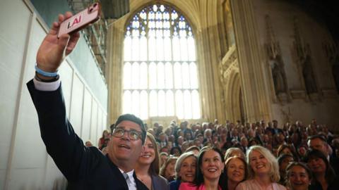 Andy Burnham holds up his phone to take a selfie with grinning Labour MPs on the steps of Westminster Hall, with the sun streaming down on them through a large window