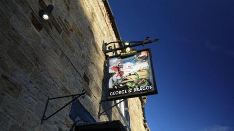 A pub sign hanging from a stone wall. The colourful sign includes a knight on horseback and a dragon reflecting the pub's name - George & Dragon.