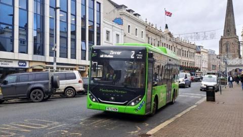 A green single decker bus parked at a bus stop in Hereford. The road has parked cars to one side with shops and office buildings to the left. In the background is a church and people walking down the nearby pavement.