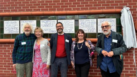 Five members of the families of organ donors line up along the ceramic mural to smile for a photograph.