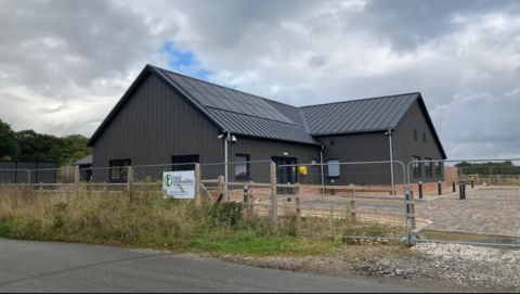 A new community centre. It is a modern building with a pitched roof. It is behind fencing. There is a paved area outside the building.