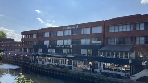 A dated riverside building with a red-brick façade and a “Premier Inn” sign above rows of windows. At ground level, a pub restaurant stretches along the water with outdoor seating and globe lights. A walkway runs beside the river where a few people stand, while the calm water reflects the structure under a clear sky.