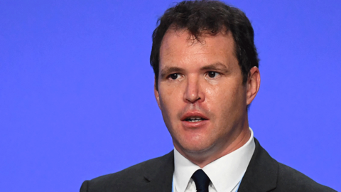 Lee Waters is standing on a stage delivering a speech. He has brown hair, is cleanly shaven. He's wearing a dark coloured suit and tie and a white shirt.