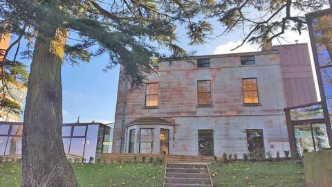 The rear view of a sandstone mansion in Dumfries viewed through a canopy of trees on a clear blue skied day