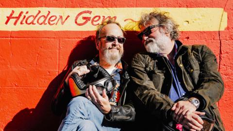 Dave Myers and Si King sit in front of a painted orange wall. They are both wearing biker jackets and have sunglasses on. Dave is smiling contentedly and Si is looking at Dave with fondness. Behind them, written on the wall, is a sign that reads "Hidden Gem".