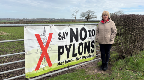 Kath Hardman wearing beige coat stood next to a farmers fence with a "No to pylons" sign on it.