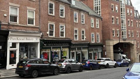 The LD Mountain Centre in Dean Street, Newcastle. The building has a shop occupying the ground floor with two floors above. Cars are parked in a row outside and Pure Knead Bakery is next door on the left.