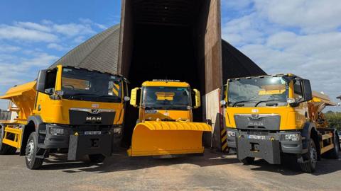 Two yellow and black gritter machines parked either side of a yellow snow plough outside a large domed grit store