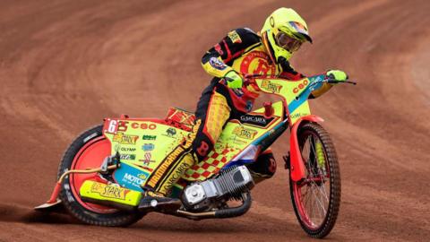 A person riding a speedway motorbike on a brown dirt track. The bike is yellow with red paint and dozens of sponsor logos over the body of it. The rider is wearing a red, black and yellow race suit with a yellow helmet and gloves