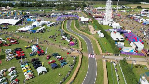 An aerial view of the festival in green fields, with parked cars on the left, a racing track running through the middle and a large white ferris wheel behind and to the right,