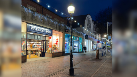 The image shows a high street with lit up shops like Waterstones and Superdrug. In the centre of the image is a streetlight that has Christmas lights hanging from it. A white sheet has been stuck to the neck of the structure.